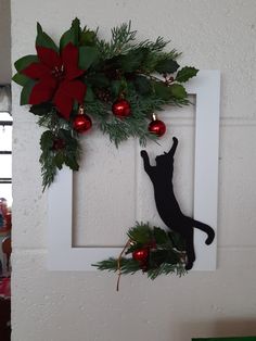 a black cat sitting on top of a white frame next to a christmas wreath and ornaments