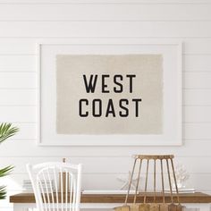 a white dining room table with two chairs and a framed west coast sign on the wall