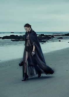 a woman is walking on the beach wearing a long black dress with sheer layers and gloves