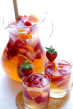 two glasses filled with liquid and strawberries next to pitcher of lemonade on a wooden tray