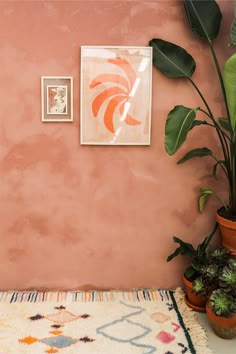 two potted plants sit on a rug in front of a pink wall with an orange and white painting