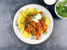 a white plate topped with pasta and meat covered in sauce next to a bowl of sour cream