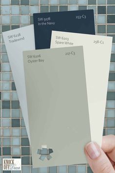 a hand holding three different shades of gray, white and grey paint swatches in front of a mosaic tile wall