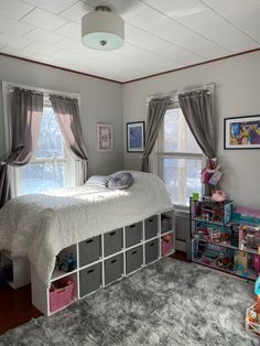 a bed room with a neatly made bed and lots of toys on the floor next to two windows