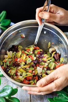 two hands holding a metal bowl filled with chopped vegetables and olives on top of a wooden table