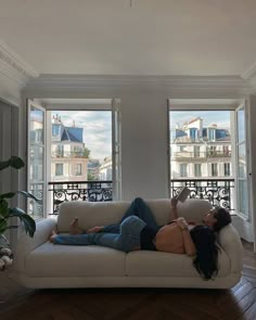 a woman laying on top of a white couch in front of two large windows next to a wooden floor