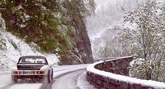 a car driving down a snow covered road next to a mountain side with trees on both sides