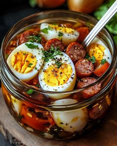 a glass bowl filled with hard boiled eggs, tomatoes and sausages on top of a wooden cutting board