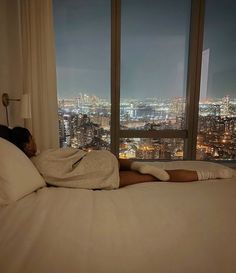 a person laying on a bed in front of a window with the city lights lit up