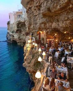 people are sitting at tables on the cliff by the water and eating food in front of them