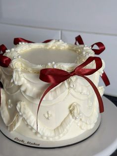 a white wedding cake with red ribbon and bow on top, sitting on a plate