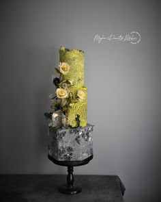 a three tiered cake decorated with flowers and lace on a black stand against a gray wall