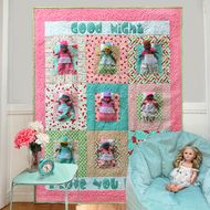 a doll sitting on a blue chair in front of a quilted wall hanging from the ceiling