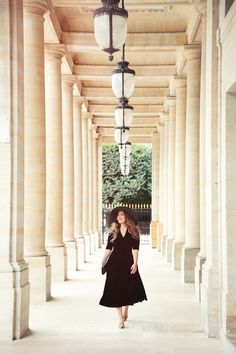 a woman in a black dress and hat walking down an empty walkway with columns on both sides