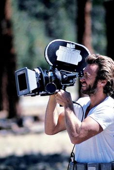 a man holding a camera up to his face with trees in the back ground behind him