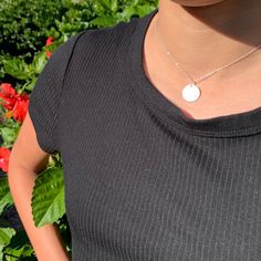 a woman standing in front of some flowers wearing a black shirt and silver necklace with a white disc on it