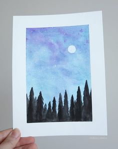 a hand holding up a card with trees and the moon in the sky behind it