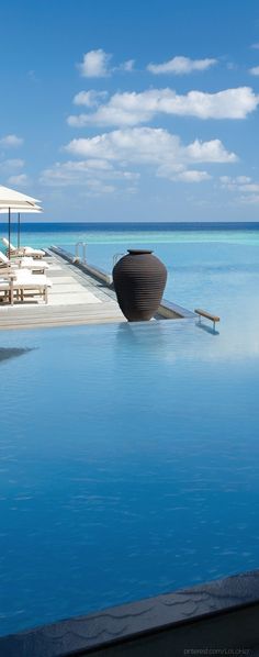 an empty swimming pool with chairs and umbrellas on the edge, in front of blue water