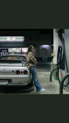 a woman standing next to a silver car at a gas station with her hand on the fuel pump