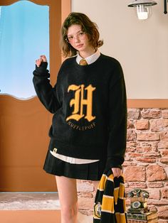 a woman standing in front of a brick wall wearing a black sweater with the letter h on it