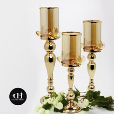 three gold colored candlesticks with flowers on the table next to them and a white background