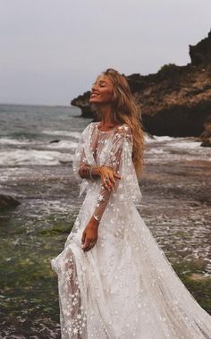 a woman standing on the beach wearing a white dress with long sleeves and sheer lace