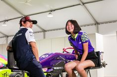 a man standing next to a woman sitting on top of a purple race car in a garage