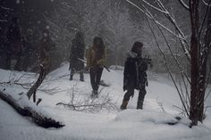 a group of people walking through the snow in front of trees and bushes with one person holding a knife