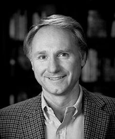 a black and white photo of a man in a suit smiling at the camera with bookshelves behind him