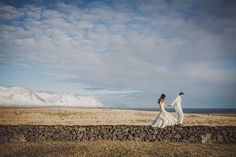 a man and woman holding hands walking across a dry grass field with mountains in the background