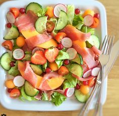 a white plate topped with cucumbers, tomatoes and other vegetables next to silverware