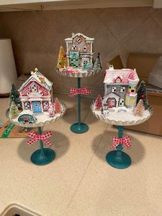 three small christmas houses on pedestals sitting on a kitchen counter with boxes in the background