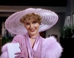 a woman in a pink dress and hat holding a binder with her right hand