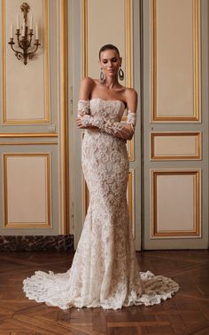 a woman standing in front of a wall wearing a wedding dress with long sleeves and an off the shoulder design