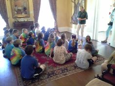 a group of children sitting on the floor in front of a woman talking to them