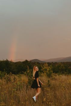 a woman in a black dress walking through tall grass with a rainbow in the background