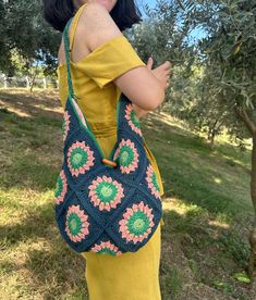 a woman holding onto a crocheted bag in an olive tree field with grass and trees behind her