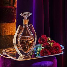 a glass decanter on a tray with red flowers in front of purple curtains