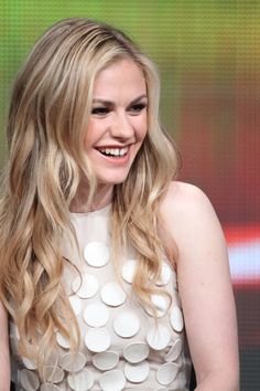 a woman with long blonde hair smiles while sitting in front of a green screen wearing a white dress