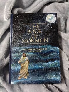 the book of mormon is laying on a bed with grey sheets and a moon in the background