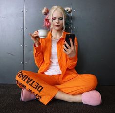 a woman in an orange prison outfit sitting on the floor holding a cup and cell phone