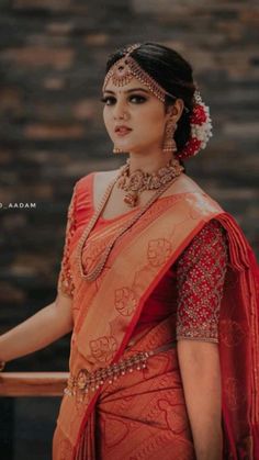 a woman in an orange and red sari with jewelry on her head, standing next to a brick wall