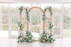 an arch decorated with flowers and greenery stands in front of two large open windows