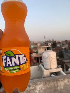 a person holding up a bottle of fanta orange flavored drink in front of a cityscape