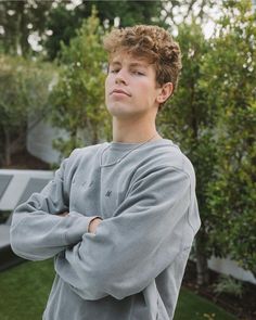 a young man with his arms crossed standing in front of some bushes and trees,