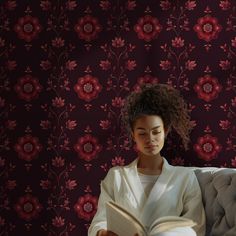 a woman sitting on a couch reading a book in front of a floral wallpaper