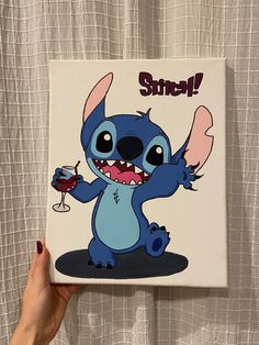 a hand holding up a small square painting with stitch on it's face and mouth