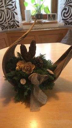 a wooden table topped with a bowl filled with pine cones and greenery next to a window