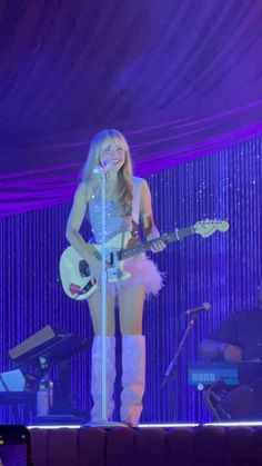 a woman standing on top of a stage with a guitar in her hand and singing into a microphone