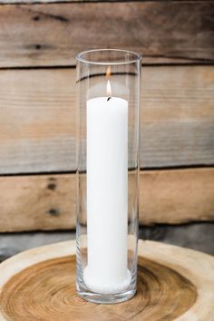 a white candle is sitting in a glass vase on a wooden table next to a wood slice